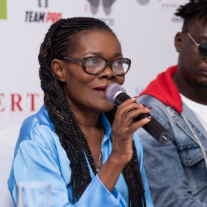 Suzanne Dimosi de face, micro à la main, des lunettes sur le nez, des tresses et un chemisier bleu satiné